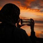 Rachunek sumienia na co dzień: prosta praktyka dla zabieganych Kobieta fotografuje zachód słońca nad jeziorem na tle gór